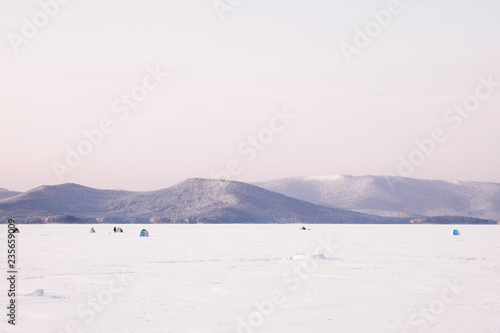 Landscape with fishermen and tilts tents on winter lake covered with snow and ice. Winter extreme sports - ice fishing.