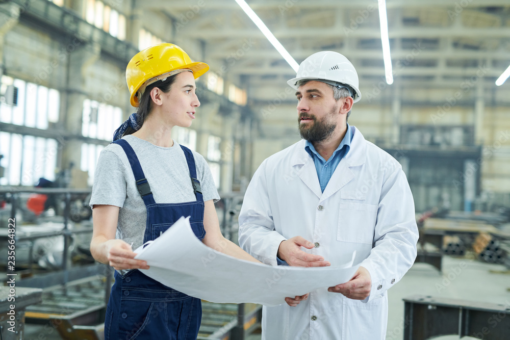 Waist up portrait of female factory worker discussing plans and blueprints with mature engineer in workshop Waist up portrait of female factory worker discussing plans and blueprints with mature engineer in workshop