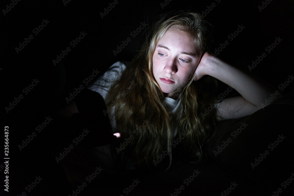 Depressed girl lying down on a couch in the dark while using her smartphone. The light from the screen is illuminating her face. Depressed girl lying down on a couch in the dark while using her smartphone. The light from the screen is illuminating her face.