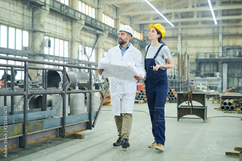 Full length portrait of female factory worker discussing plans with mature engineer walking in workshop, copy space