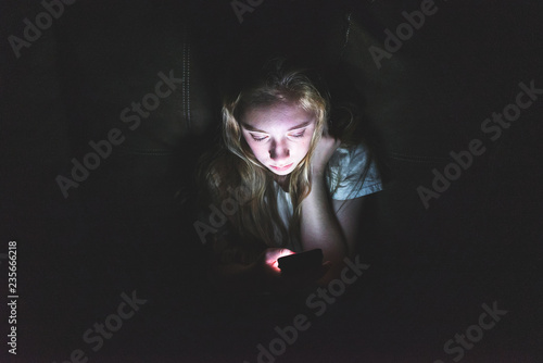Upset girl sitting in the dark while using her smartphone. The light from the screen is illuminating her face.