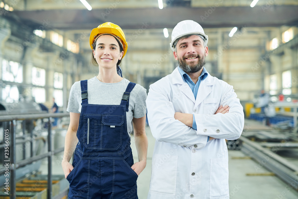 Waist up portrait of two smiling factory workers looking at camera while posing in industrial workshop Waist up portrait of two smiling factory workers looking at camera while posing in industrial workshop