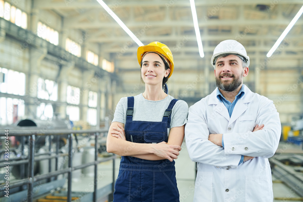 Waist up portrait of two smiling factory workers standing with arms crossed and looking away while posing in industrial workshop, copy space Waist up portrait of two smiling factory workers standing with arms crossed and looking away while posing in industrial workshop, copy space