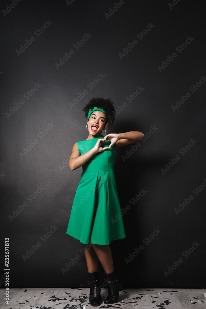 Full length photo of young afro american lady wearing green dress showing heart shape with fingers, isolated over dark background Full length photo of young afro american lady wearing green dress showing heart shape with fingers, isolated over dark background
