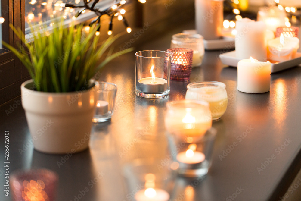 hygge, decoration and christmas concept - candles burning in lanterns and festive garland on window sill at home