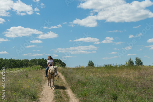 Photo of the small woman on a big horse in summer field. Image of happy female sitting on purebred horse.