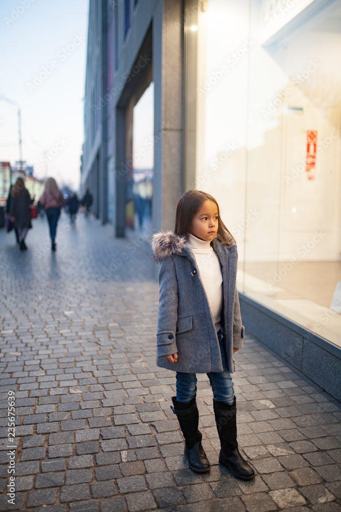 Child girl is standing next to shopwindow on city street in evening.