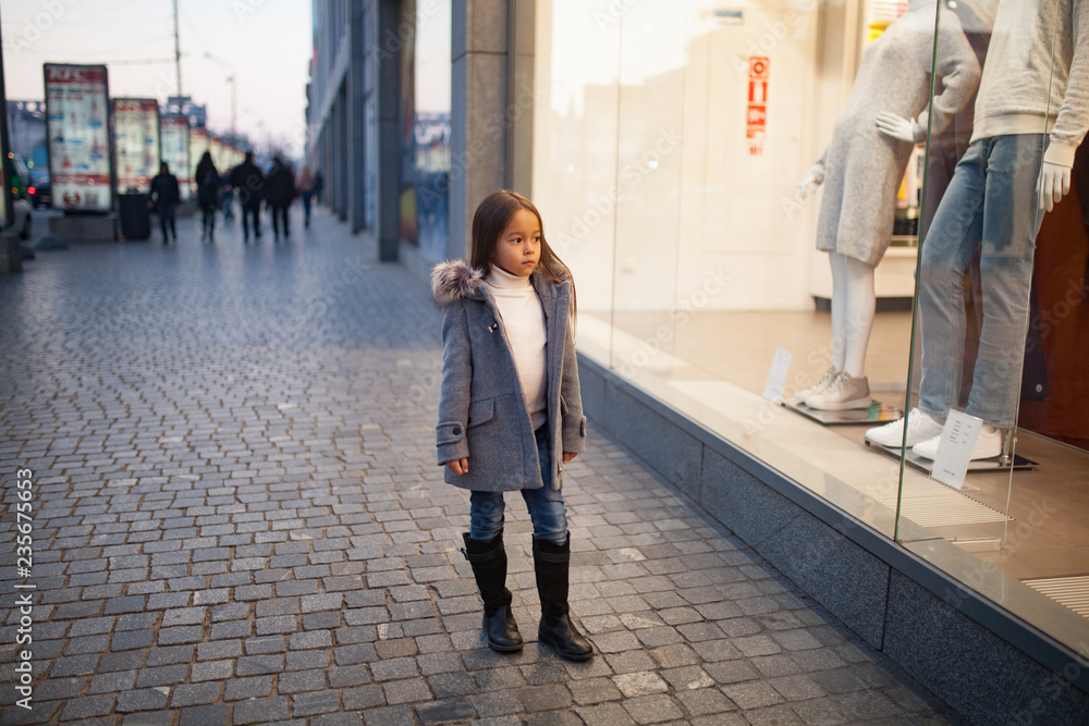 Child girl is standing next to shopwindow on city street in evening ...
