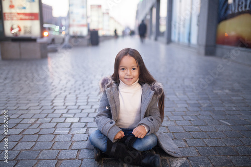 Child girl is sitting and smiling in the middle of the sidewalk in evening.