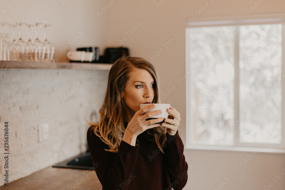 Pretty Young Blonde Woman Sipping Morning Hot Coffee in Modern Designed Kitchen While Looking out Bright Interior Window in the Winter Pretty Young Blonde Woman Sipping Morning Hot Coffee in Modern Designed Kitchen While Looking out Bright Interior Window in the Winter