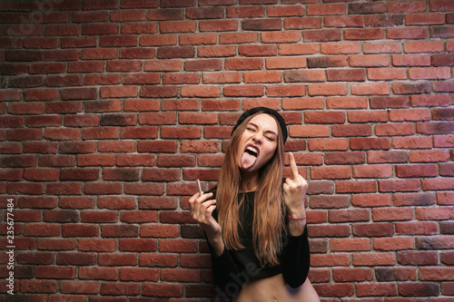 Image of cheeky sporty girl, standing against brick wall with cigarette and showing fuck you gesture