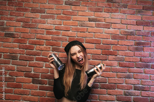 Image of subcultural hip hop girl 20s, standing against brick wall with spray cans photo