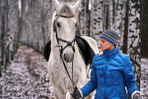 Young pretty woman in a blue jacket and a sports hat for a walk with a white horse in the winter birch forest.