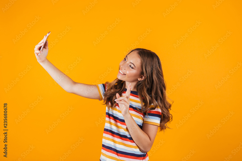Happy young girl posing over yellow wall background take a selfie by mobile phone. Happy young girl posing over yellow wall background take a selfie by mobile phone.