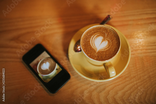 Cup of coffee with a heart in the foam, with a mobile phone next to it.