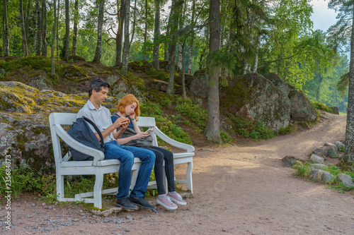 Handsome middle-aged man and young pretty lady sitting on bench and using smartphones in summer evening. Tourists on the beautiful landscape background. Monrepos Park, Vyborg, Russia. Travel concept