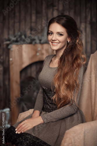Close up portrait of young cheerful beautiful girl with dark long curly hair in gray dress in cozy interior. Brunette smiling with teeth, looking in camera with happy and relaxed face expression.