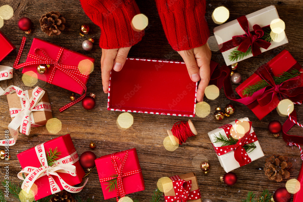 Christmas gift giving - hands preparing and wrapping red and white paper christmas gift boxes on wooden background with bokeh lights