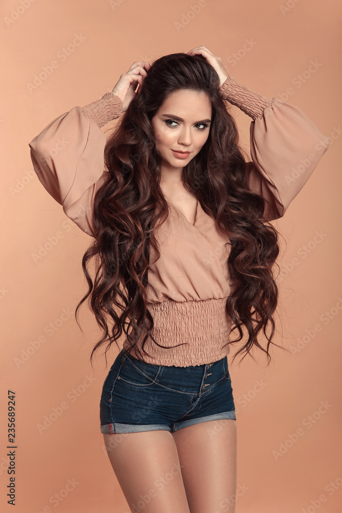 Fashionable beautiful girl wearing blouse with long sleeves and denim  shorts isolated on beige studio background. Sensual brunette with makeup  and long healthy curly hair style posing in trendy outfit Stock Photo |