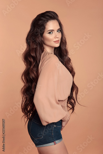 Fashionable beautiful girl wearing blouse with long sleeves and denim  shorts isolated on beige studio background. Sensual brunette with makeup  and long healthy curly hair style posing in trendy outfit Stock Photo |
