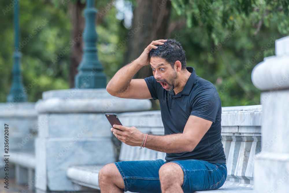 Man holding a smart phone with surprised expression in a park. Emotion and feeling concept Man holding a smart phone with surprised expression in a park. Emotion and feeling concept