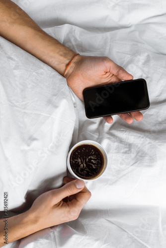 partial top view of man holding cup of coffee and smartphone with blank screen in bed