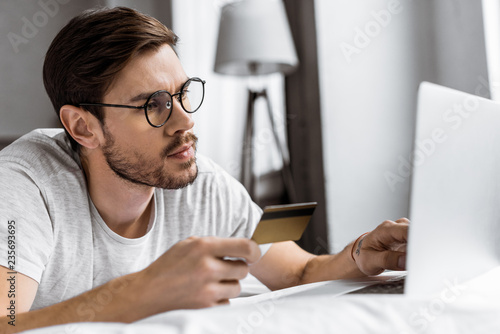 handsome young man in eyeglasses holding credit card and using laptop on bed