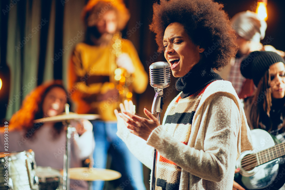 Fototapeta premium Close up of mixed race woman singing. In background band playing instruments. Home studio interior.