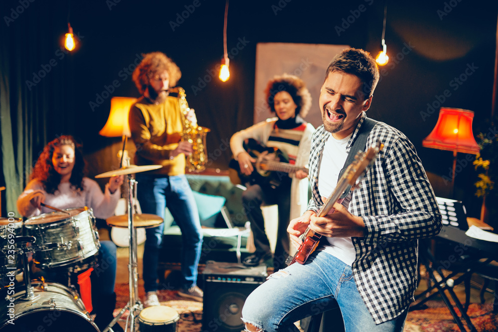 Naklejka premium Man singing and playing bass guitar while sitting in home studio. In background his band playing instruments.