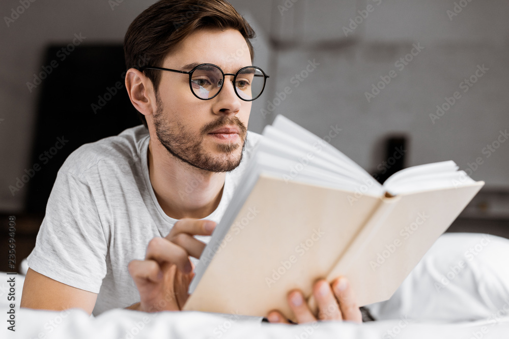 handsome young man in eyeglasses lying in bed and reading book handsome young man in eyeglasses lying in bed and reading book