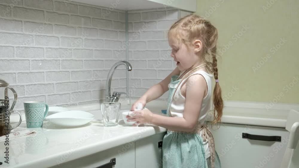 beautiful little girl in an apron is standing on a chair, in the bright kitchen, doing the dishes with a sponge, helping parents beautiful little girl in an apron is standing on a chair, in the bright kitchen, doing the dishes with a sponge, helping parents
