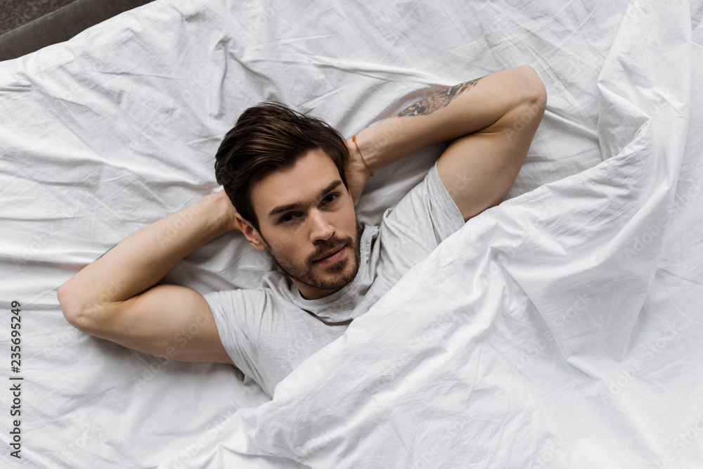 top view of attractive young man lying under blanket in bed and looking at camera top view of attractive young man lying under blanket in bed and looking at camera