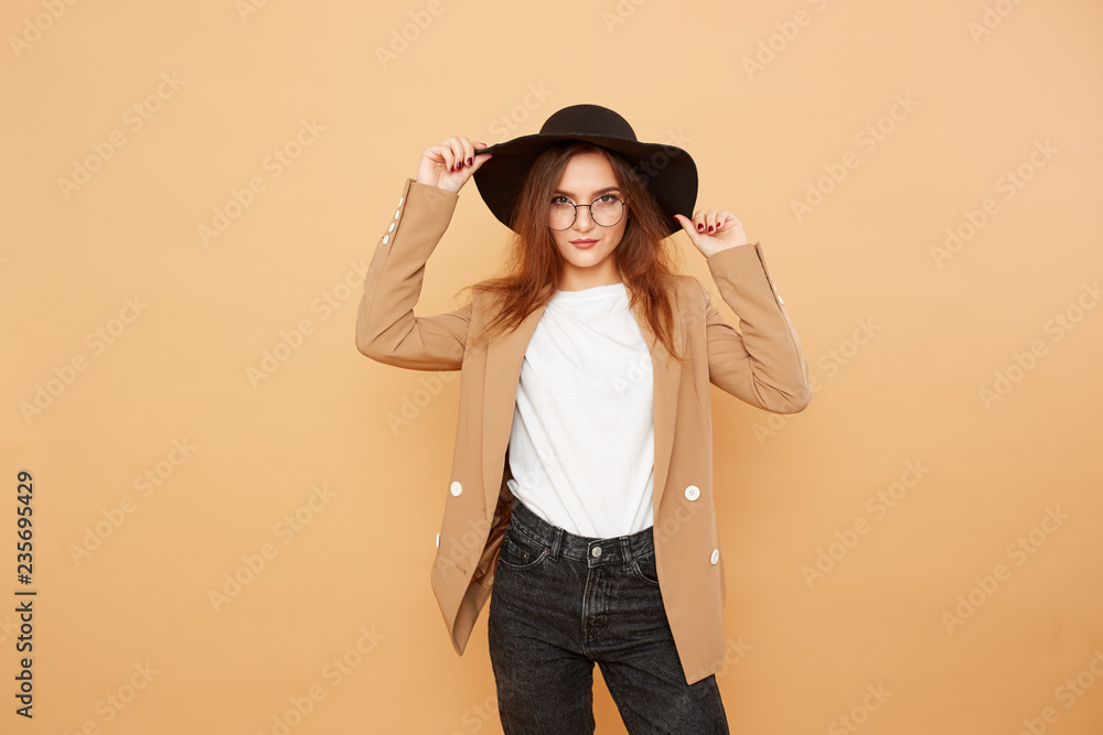 Pretty brunette girl with long hair in glasses and a black hat on her head dressed in white t-shirt, jeans and beige jacket is posing on the beige background in the studio Pretty brunette girl with long hair in glasses and a black hat on her head dressed in white t-shirt, jeans and beige jacket is posing on the beige background in the studio
