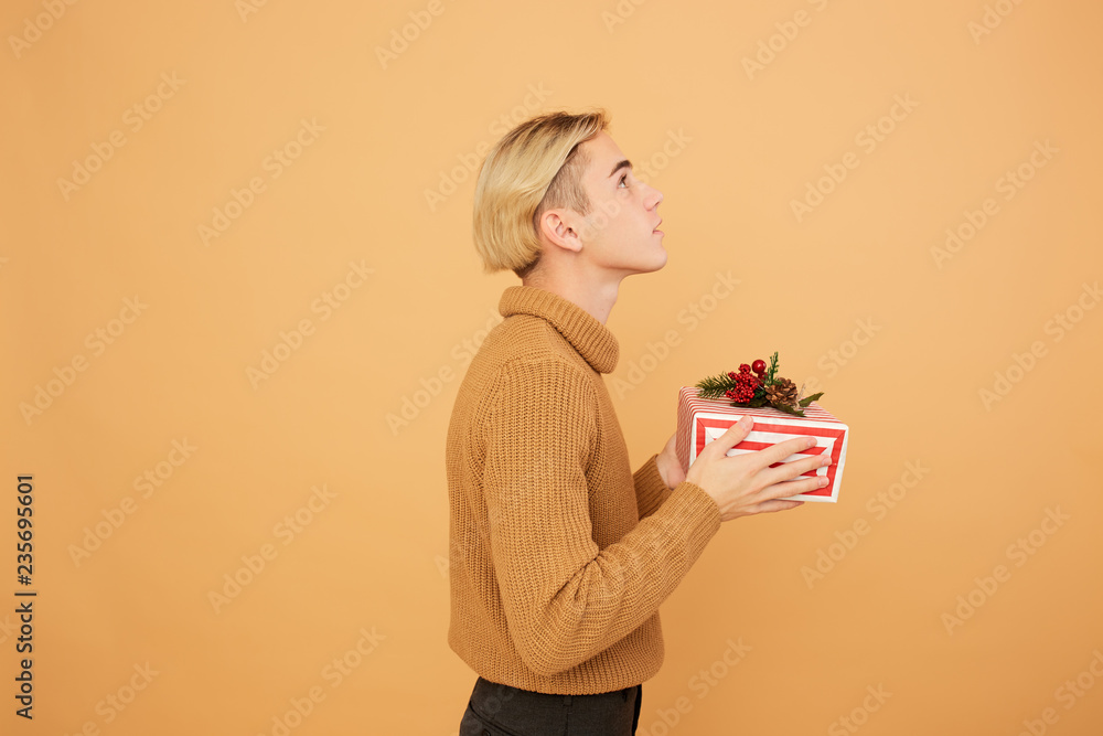 Young blond guy dressed in mustard color sweater holds a Christmas gift in his hands on the beige background in the studio Young blond guy dressed in mustard color sweater holds a Christmas gift in his hands on the beige background in the studio