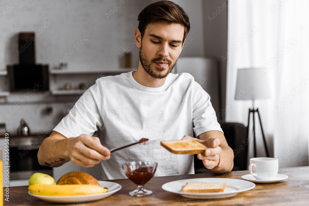 smiling young man applying jam onto toast on weekend morning at home smiling young man applying jam onto toast on weekend morning at home