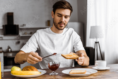 smiling young man applying jam onto toast on weekend morning at home