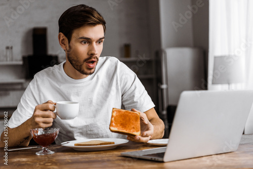shocked young freelancer eating toast with jam and looking at laptop screen at home