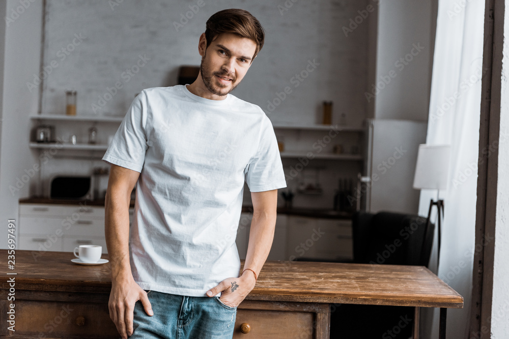 smiling young man leaning back on kitchen table and looking at camera at home