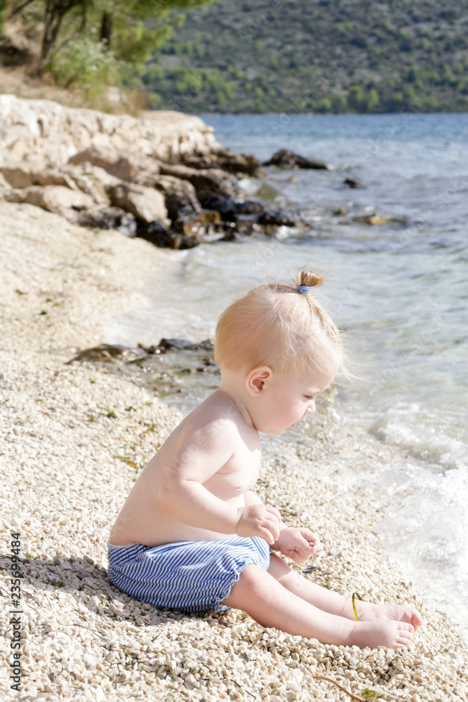Candid photo of adorable blonde baby on the pebble beach near the sea, summer vacation on the seaside Candid photo of adorable blonde baby on the pebble beach near the sea, summer vacation on the seaside
