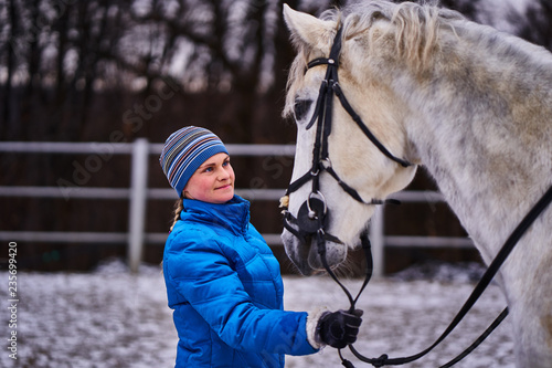 Young pretty woman in blue jacket and sports hat on a walk with a white horse on a winter cloudy day. A horse licks its hands to its owner.