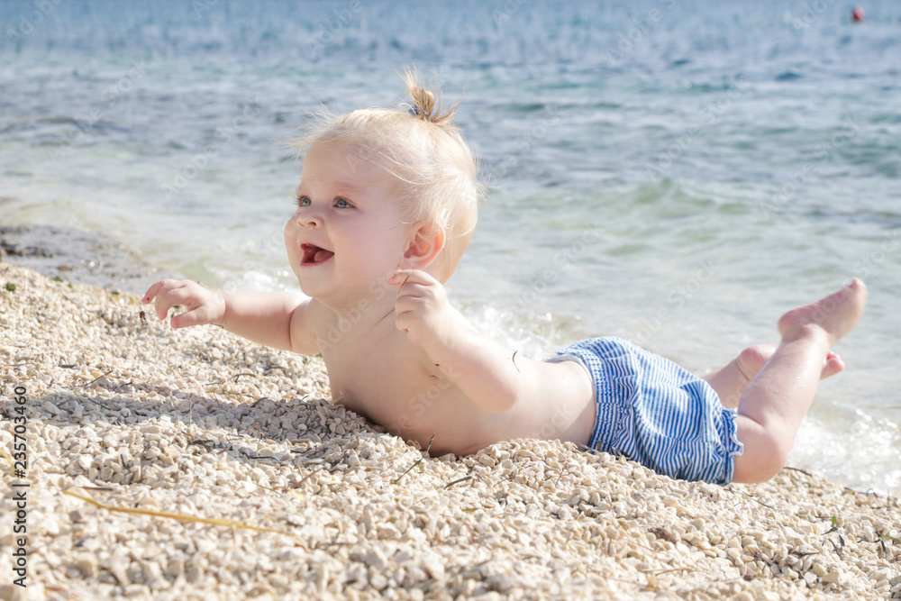 Candid photo of adorable blonde baby on the pebble beach near the sea, summer vacation on the seaside Candid photo of adorable blonde baby on the pebble beach near the sea, summer vacation on the seaside