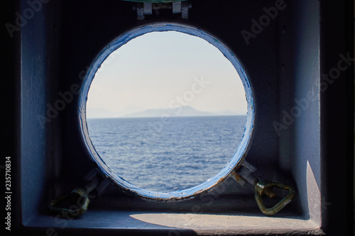 View though a ship window porthole of a traveling ferry onto the aegean sea with a small island on the horizon.