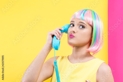 Young woman in a colorful wig talking on a retro phone on a split yellow and pink background