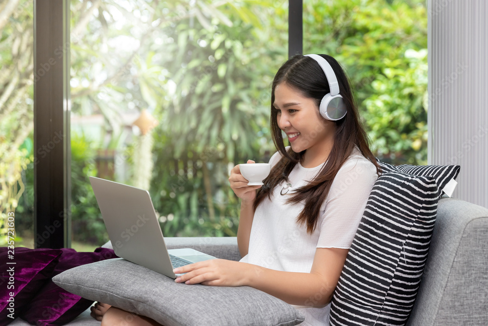 Pretty smiling young woman wear headphones sitting on sofa holding a cup of coffee and usinging on tablet. Pretty smiling young woman wear headphones sitting on sofa holding a cup of coffee and usinging on tablet.