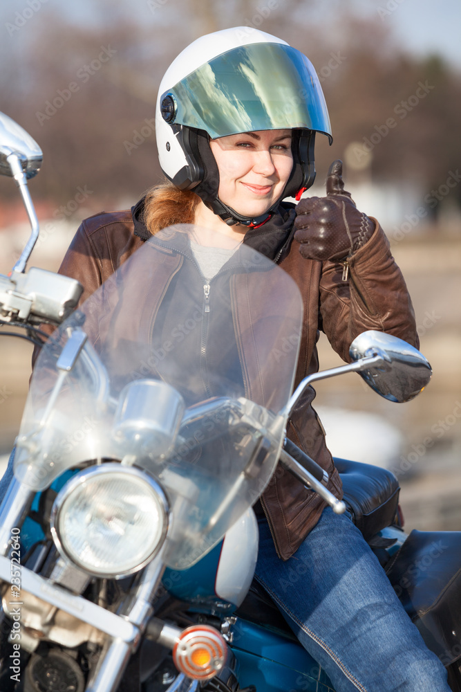 Woman motorcyclist in brown leather jacket and white crash helmet showing thumb up sign with hand Woman motorcyclist in brown leather jacket and white crash helmet showing thumb up sign with hand