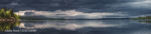 Wide angle panorama dramatic scenery landscape with lake water reflecting clouds, island with trees in sunlight, mountain range with forest in far horizon in north Russia