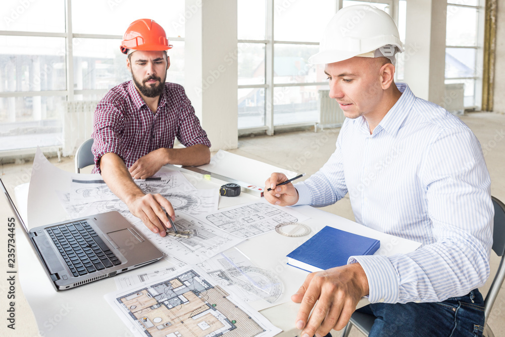 male engineers, architects working at the desk in helmets. Drawings, laptop, roulette on the desktop. Reception and supervision of building construction male engineers, architects working at the desk in helmets. Drawings, laptop, roulette on the desktop. Reception and supervision of building construction
