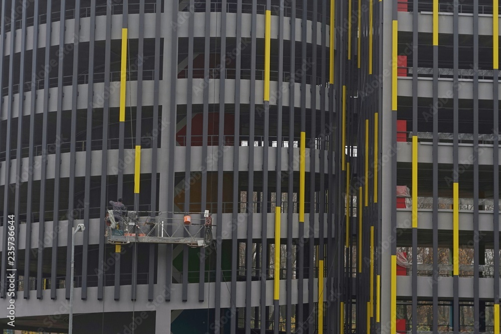 Obraz premium the builder is on the cradle, paints the metal constructions in yellow on the parking facade