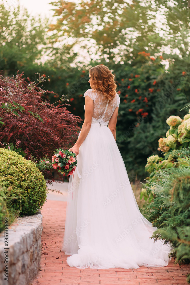young blonde girl in an elegant long white wedding light dress with a long train is standing back to the camera in a green garden, holding a lovely bouquet of pink flowers. no face art photo