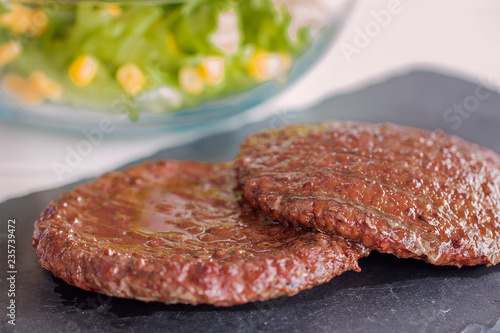 Two hamburgers. Background: a glass bowl with lettuce and mais.
